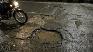 Primer plano de una carretera urbana con baches, grietas y pavimento irregular mientras una motocicleta circula cerca